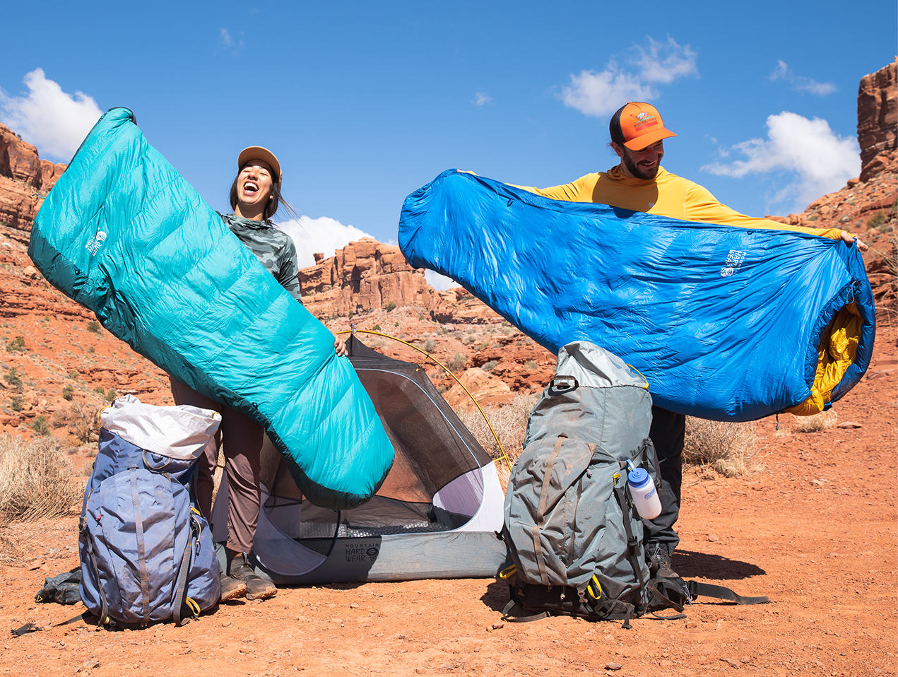 Two backpackers getting their Bishop Pass sleeping bags out of their PCT packs, stretched out bags arms wide with big smiles in the Utah Desert