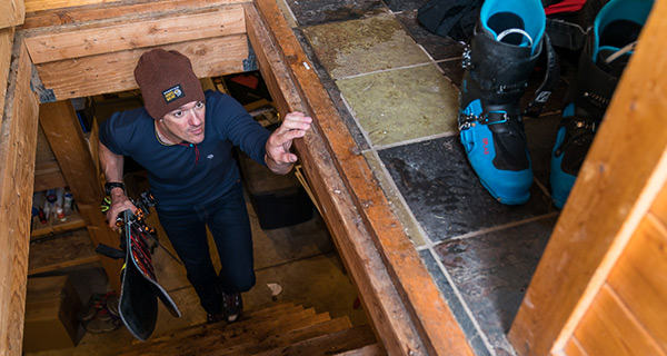 Mountain Hardwear Athlete Luke Smithwick walks upstairs in a ski hut, with old skis in hand.