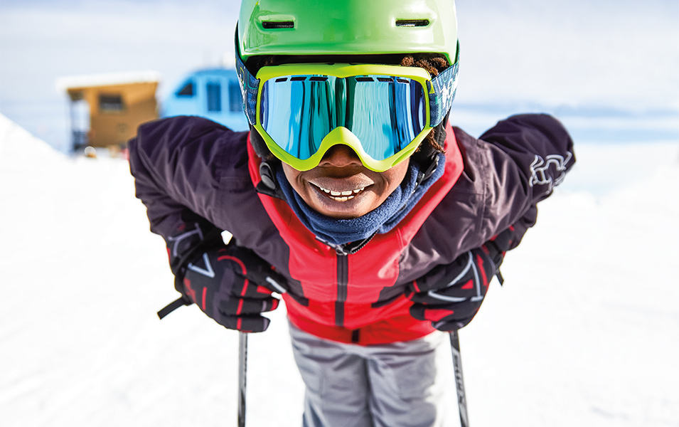 A young skier leans forward on his ski poles and smiles at the camera. 