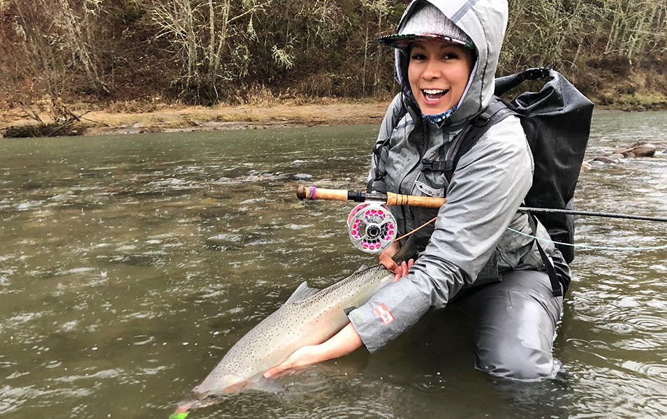 Pro angler Ruth Sims wears a rain jacket and pants while crouching down in a river holding a fish for the camera.