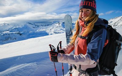 A skier stands in front of a giant cornice of untouched powder as she looks at the camera and smiles.