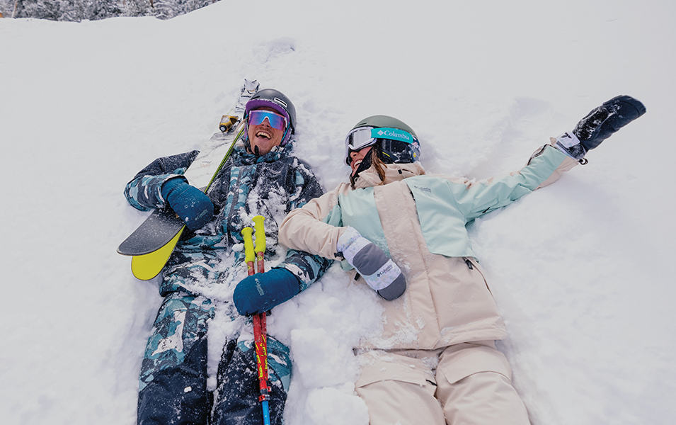 Two skiers lie on their backs in a field of snow as hey smile and laugh together. 