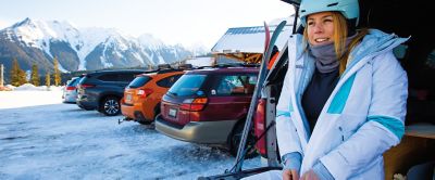 A woman stands in the parking lot of a ski resort zipping up her jacket.  