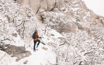 A bundled-up man in an orange puffy jacket and black snow pants stands with trekking poles on a snowy cliff watching the scenery.