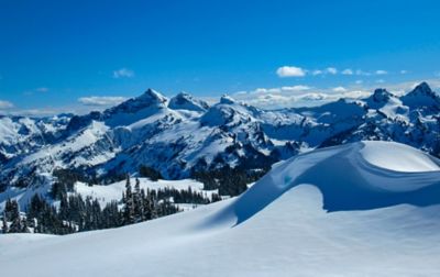 A snow-covered valley full of deep powder sits in the foreground of Mount Rainier National Park while gorgeous mountains sit in the background under a bright blue sky filled with fluffy white clouds.