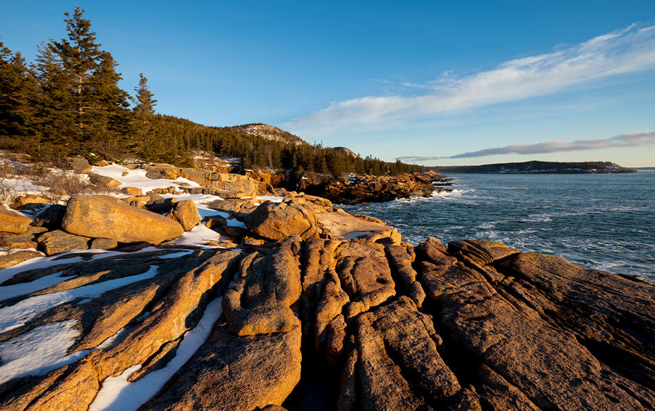 Golden rocks covered with pockets of snow sit next to the deep blue ocean as the morning light hits them in Acadia National Park.