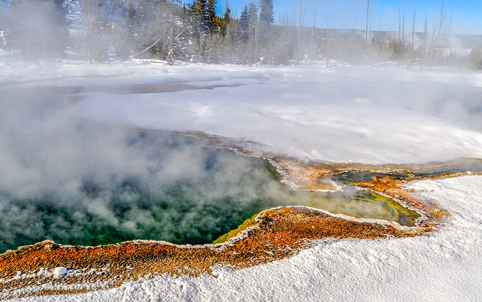 A misty geyser with green and yellow water that’s surrounded by snow emits steam in Yellowstone National Park.