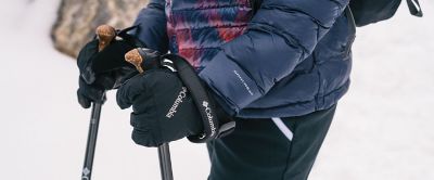 A pair of black Columbia Sportswear gloves grasp a set of trekking poles amid a wintery background. 