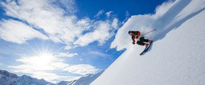 A skier comes down a steep powdery slope with sunlight in the background. 