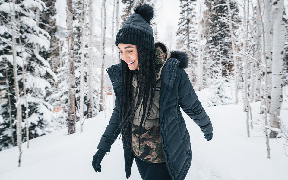 A woman with long braids wearing a black puffy jacket walks through a wintery landscape.