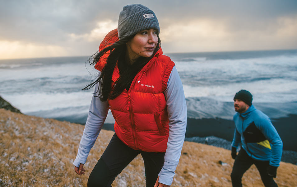 A woman in a red Columbia Sportswear vest and grey hat hikes up a frosty hillside with a man in a blue winter jacket behind her.