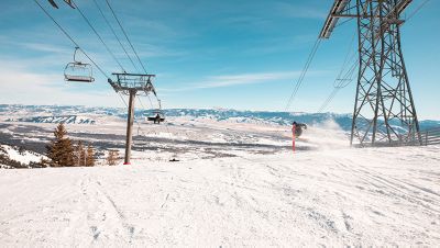 A skier mid jump on a run in a resort, in between two chair lifts
