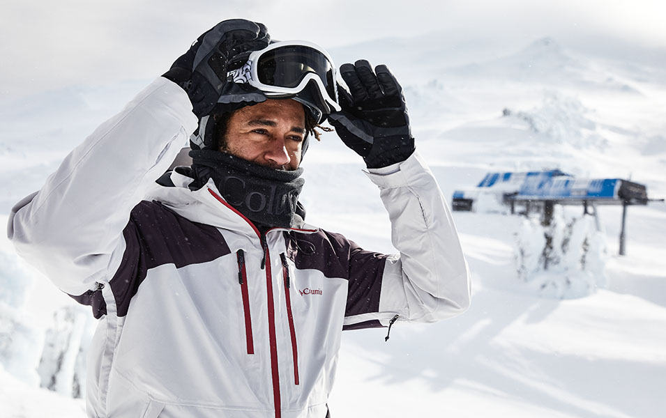 A man in a white-and-black Columbia Sportswear jacket with a red zipper adjusts his goggles as he looks into the distance while standing on a ski slope. 