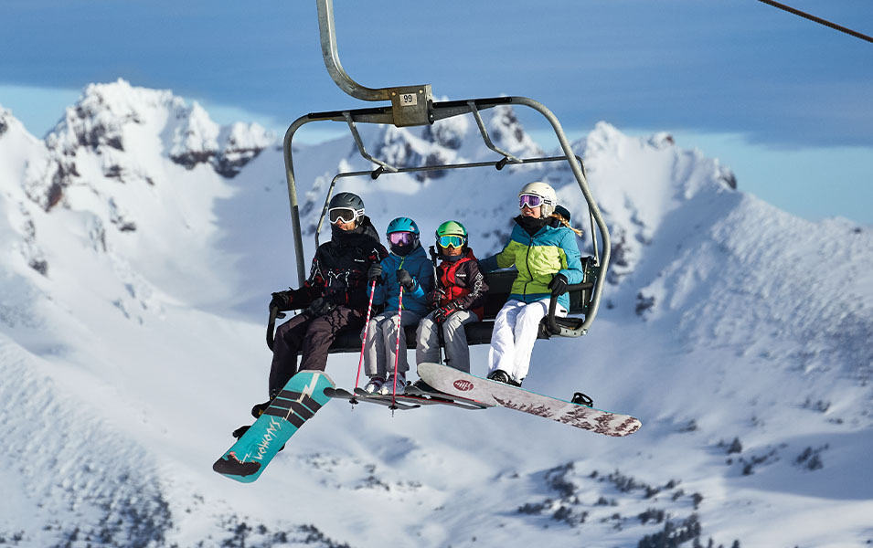  A group of skiers and snowboarders wearing Columbia Sportswear ski gear rides a chairlift with a sunny mountain range in the background.  