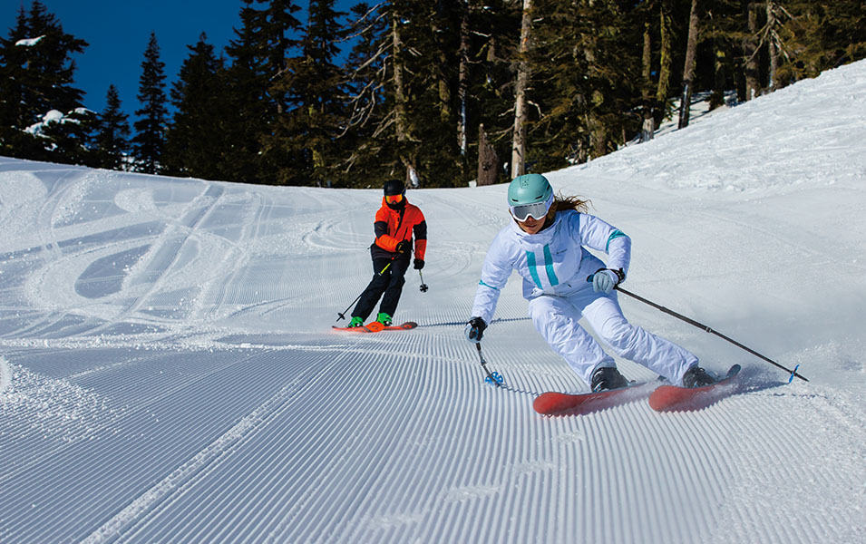A woman in a white jacket with white pants skis down freshly groomed corduroy as a man in a red jacket and black pants skis behind her. 