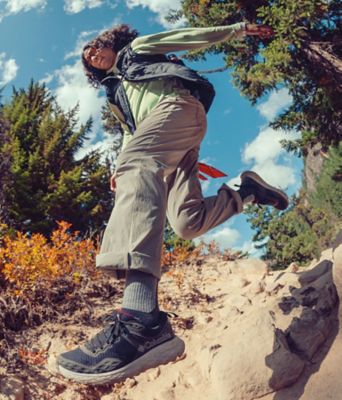 A happy man wearing a light green long sleeve shirt, grey vest, khaki pants, and Konos TRS OutDry Shoes leaps off a rock.