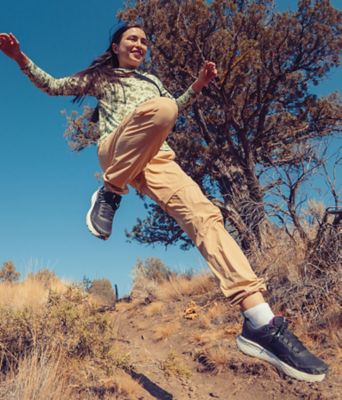 A happy women jumps off a rock wearing the Konos TRS OutDry shoe, khaki pants, and a green long sleeve shirt.