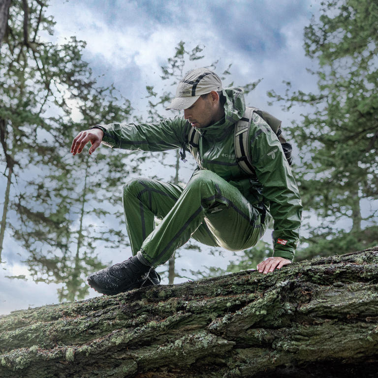 Model hopping over a large tree trunk in the green OutDry Wyldwood jacket and pants.