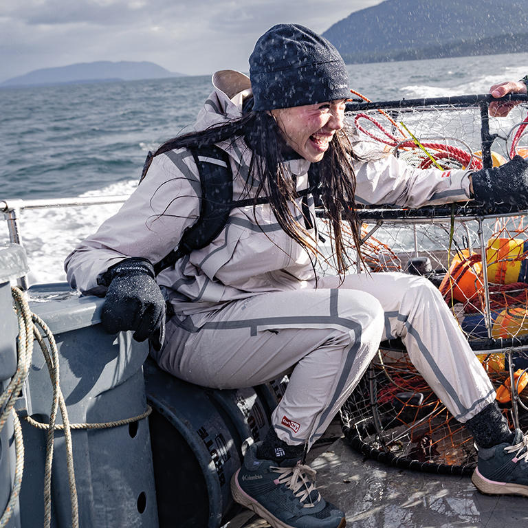 Woman on a fishing boat getting sprayed by waves and rained on with her waterproof white OutDry Wyldwood jacket and pants.