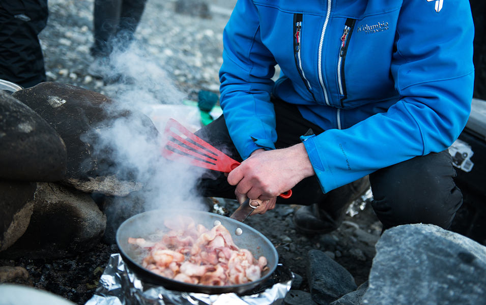 A winter camper fries bacon in a skillet over a fire. 