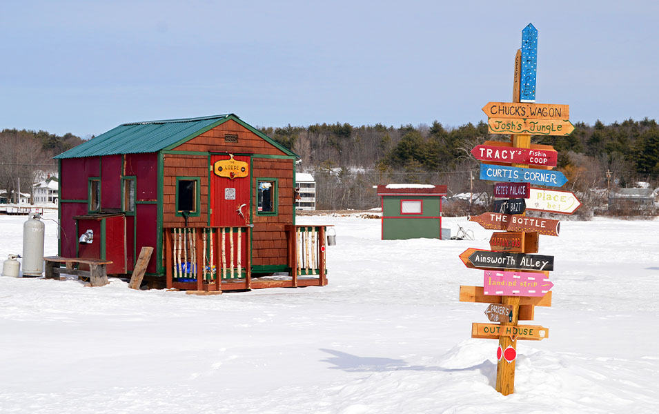 A red ice fishing hut sits on the snow in the background with a tall post containing colorful signs in the foreground.  