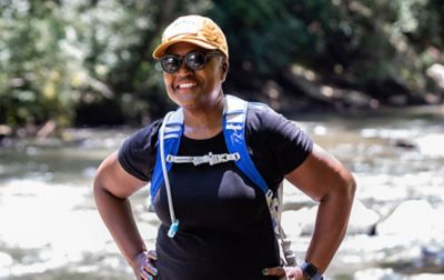 Renee Masionet, vice president of development for the Greening Youth Foundation, stands near a river wearing a hiker’s hydration pack and smiling off-camera. 
