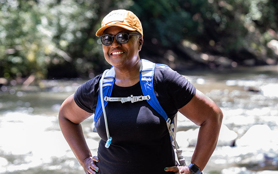 Renee Masionet, vice president of development for the Greening Youth Foundation, stands near a river wearing a hiker’s hydration pack and smiling off-camera. 