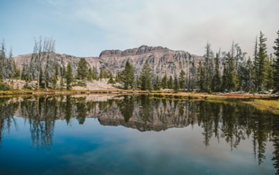 A landscape shot of a scenic blue lake with the green trees and brown rocks that stand in the background reflected in it.