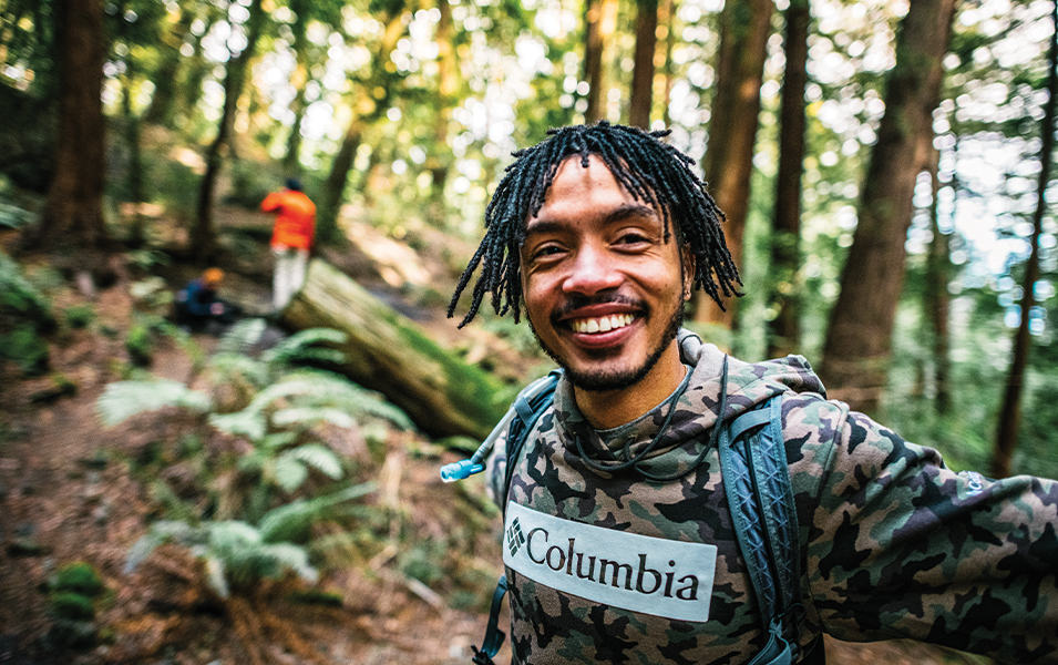 An African American man wearing a long-sleeved brown-and-green Columbia Sportswear shirt smiles widely at the camera amid a wooded forest background.