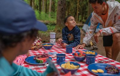 An African American family sits around a picnic table in the woods with a red-and-white checkered tablecloth and blue plates containing watermelon and other fruits.