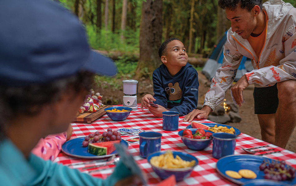 An African American family sits around a picnic table in the woods with a red-and-white checkered tablecloth and blue plates containing watermelon and other fruits.
