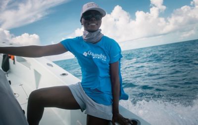 An African American woman wearing a blue Columbia Sportswear shirt smiles happily as she stands on a boat leaning against the side with waves rolling in the background.