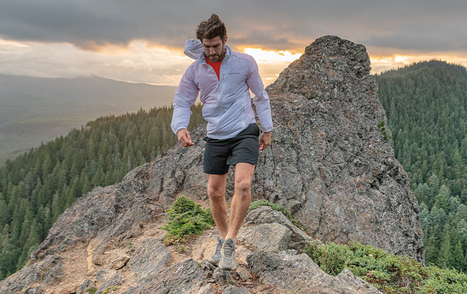 A man wearing a white Columbia Sportswear windbreaker walks along a rocky trail atop a beautiful mountain landscape. 