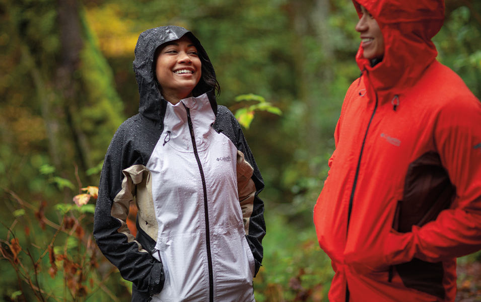 A woman stands in the rain in a forest smiling at a fellow hiker who is wearing a bright red rain coat. 