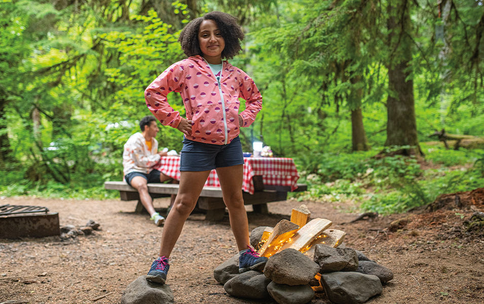 A girl wearing a pink windbreaker stands by a fire pit at a campground in a scenic forest setting. 