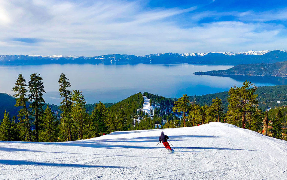 A skier in red pants heads down the slopes at Palisades Tahoe with Lake Tahoe in the background under a bright blue sky.