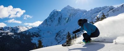 A man in a blue jacket and black helmet skis down a powdery slope with mountains in the background. 