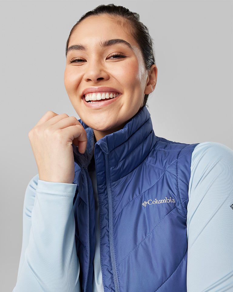 A smiling woman in a blue puffy vest and light blue long sleeve shirt.