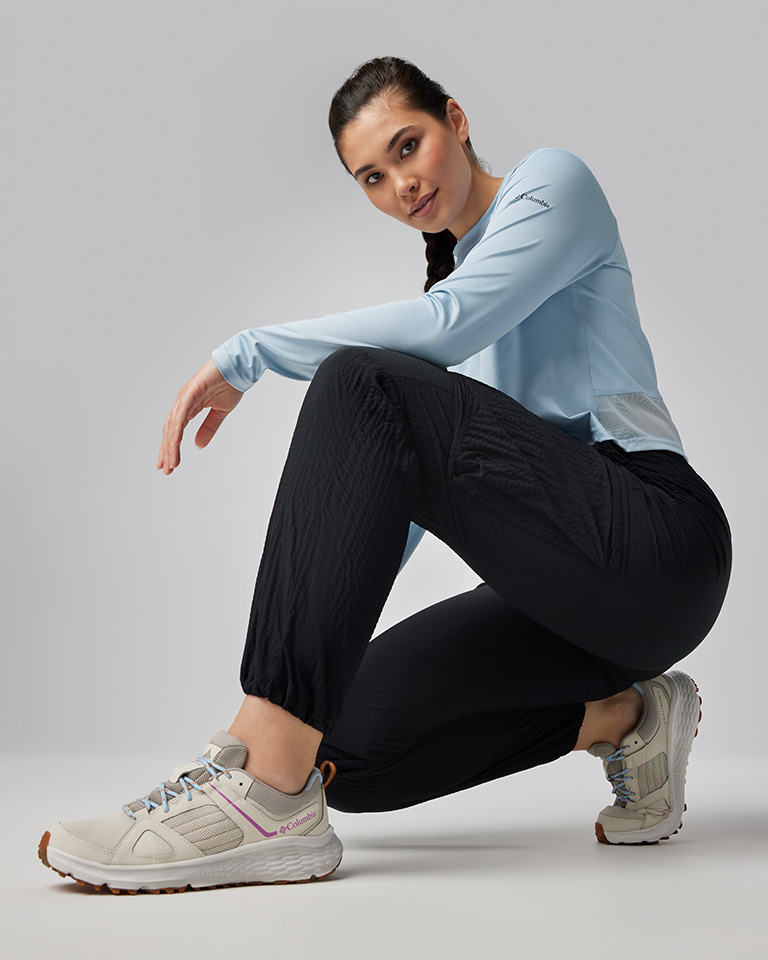 A woman kneeling in a light blue long sleeve top, black cargo pants, and light colored sneakers.