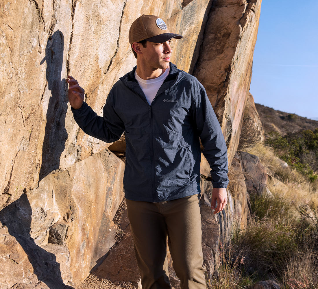 A man on a high-desert trail with a ballcap, black windbreaker jacket, white tee, and khaki pants.