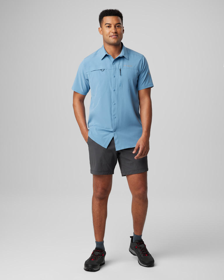A man in a blue short sleeve button up shirt and gray short with black hiking sneakers.