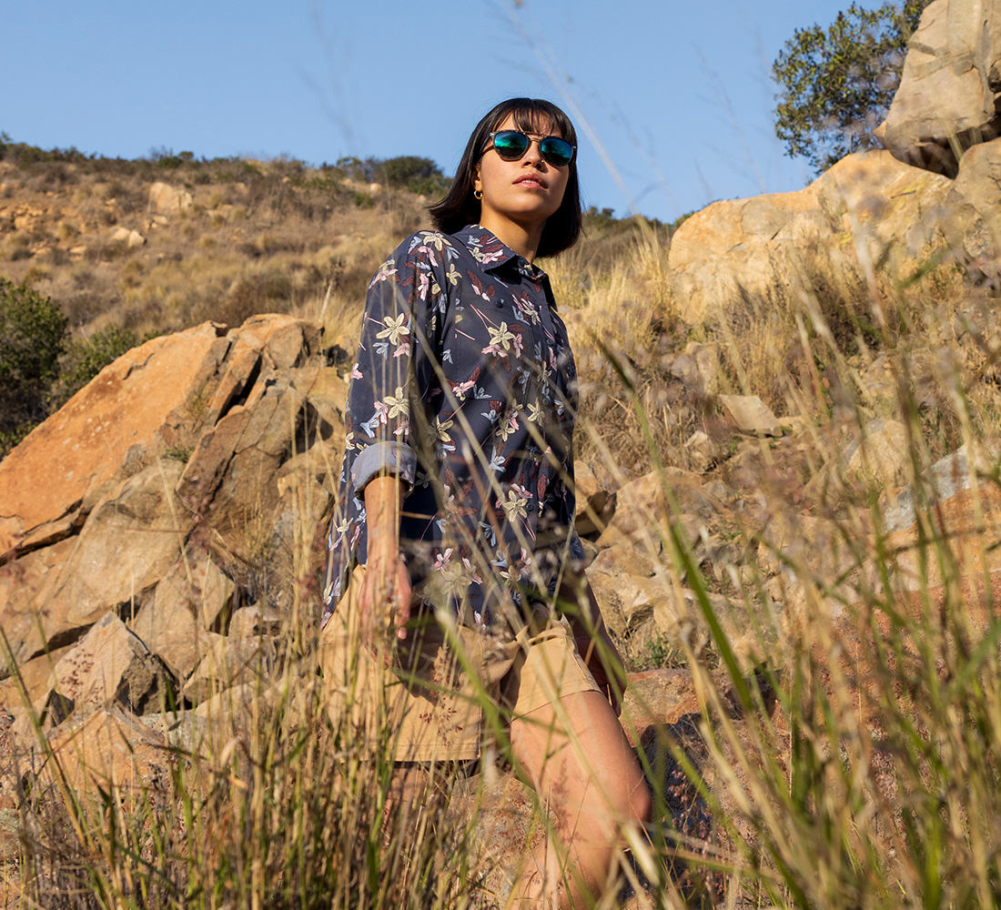 A woman in the high desert with a floral print button up, khaki shorts, and sunglasses.
