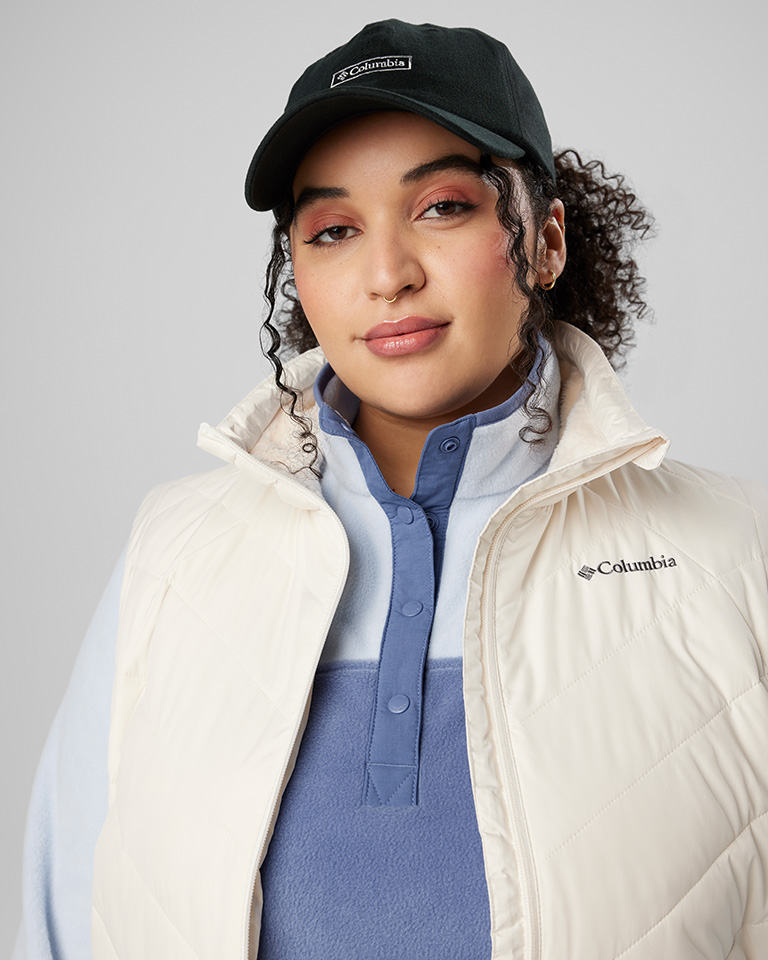 A woman in a blue fleece with a white puffy vest over the top. Plus, a black ball cap.