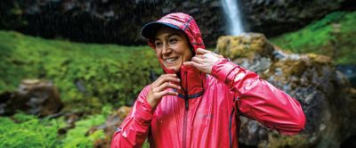 A woman wearing a bright red Columbia Sportswear rain jacket smiles widely as she reaches for her collar with green foliage and rocks in the background.