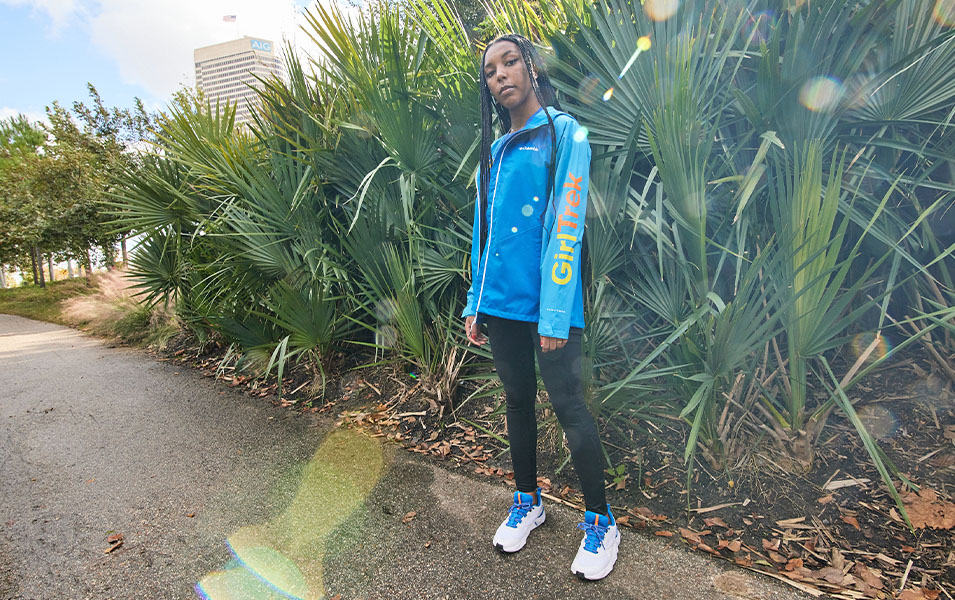 An African American woman wearing a blue GirlTrek jacket stares at the camera as she stands on a paved path with green foliage behind her. 
