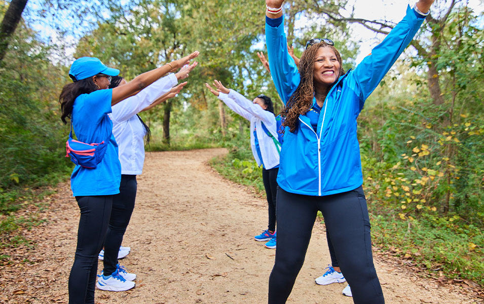 A woman in a blue GirlTrek jacket stands on a dirt trail with green foliage and throws her hands in the air enthusiastically as three woman stand behind her making a bridge. 