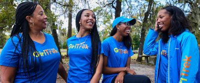 Four African American women wearing bright blue GirlTrek shirts with yellow-and-orange logos on the front smile and laugh togther in an urban park. 