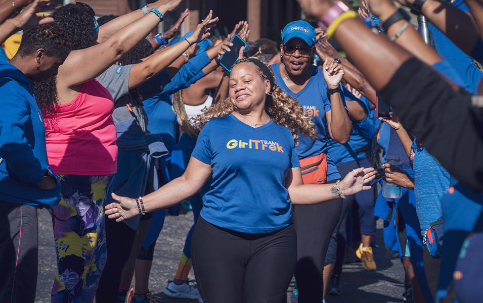 A woman in a blue GirlTrek shirt walks towards the camera enthusiastically as a group of women stand around her making a bridge. 