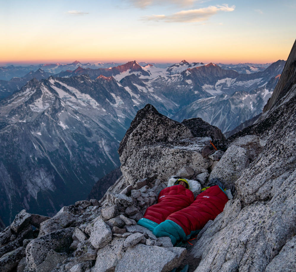 Two climbers camping cowboy style on the rocky alpine terrain as the sun rises over the mountains in the background.