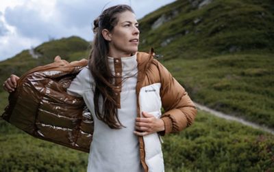 A hiker stands in front of a lush mountain trail taking off a brown Columbia Sportswear puffer coat. Underneath, she is wearing a white fleece midlayer.